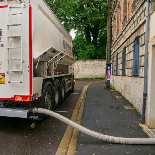 White tanker truck with a long hose trailing along the pavement, parked on a narrow street beside a stone building with blue shutters and green trees in the background.