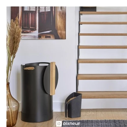 Contemporary interior with a black cylindrical trash can, wooden-handled dustpan, vase of dried pampas grass, and an open wooden staircase against a white wall.